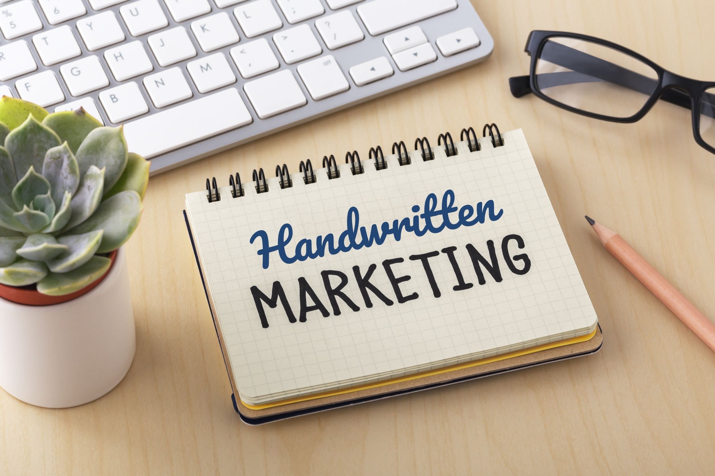 handwritten marketing written in notebook that is next to plant, glasses, pencil, and keyboard
