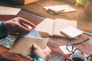 Man putting handwritten makerting letter inside an envelope. Glasses, coffee, and book on the table.